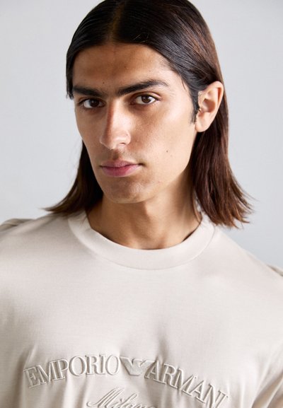 Camiseta de algodón beige con cuello redondo, que presenta el logotipo bordado de "Emporio Armani". Textura suave y diseño ajustado.