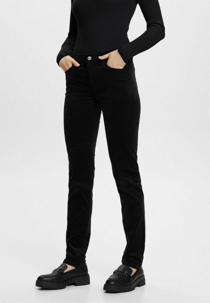 Pantalon en velours côtelé noir à coupe slim, avec une fermeture à boutons et des poches avant, assorti à des chaussures noires sans lacets. Texture lisse.