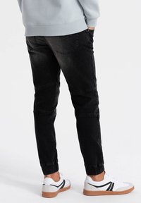 Joggers de mezclilla negra con corte ajustado, con costuras visibles, bolsillos laterales y puños elásticos, combinados con zapatillas blancas y negras.