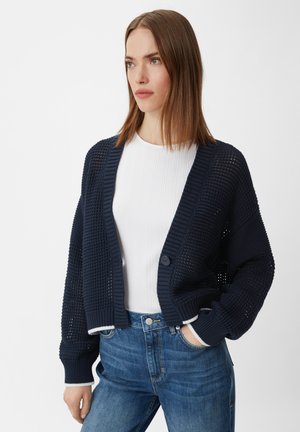 Marineblaues gestricktes Strickjacke mit offenem Frontbereich, gerippten Bündchen und einem strukturierten Muster. Kombiniert mit einem weißen, taillierten Oberteil und blauen Jeans.