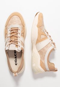Par beige och tan-färgade Coach-sneakers med strukturerat tyg, mockadetaljer och vita sulor, visade från ovan och sidor på en vit bakgrund.