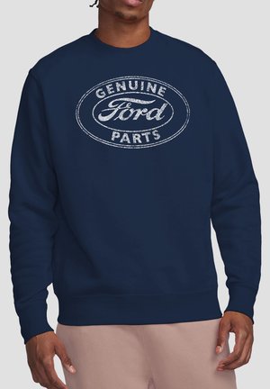Mann trägt einen marineblauen Pullover mit weißem, abgenutztem "Genuine Ford Parts"-Logo in Oval auf der Brust.