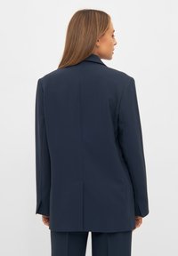 Tengerészkék zakó, strukturált kialakítással, bemetszett gallérral, hátsó egyetlen hasítékkal és egyenes szabású ujjal, amely jól megformált megjelenést kölcsönöz.