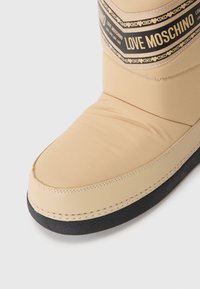 Bota bege com um cabedal liso e almofadado, sola de borracha preta e faixa castanha com "LOVE MOSCHINO" num padrão repetido.