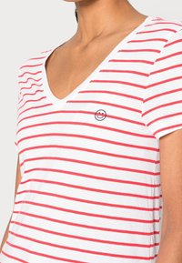 Kvinde iført hvid V-hals T-shirt med røde vandrette striber og et lille sort smiley-ansigt broderet på venstre bryst.