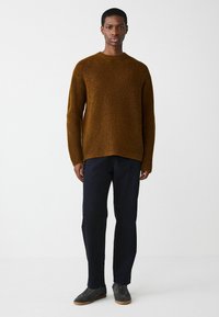 Brauner Strickpullover mit strukturiertem Muster, Rundhalsausschnitt und lässiger Passform, kombiniert mit dunklen Hosen und grauen Sneakers.
