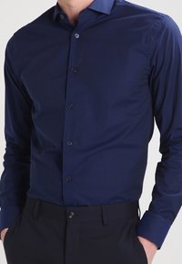 Man in een aansluitend, lang mouw, donkerblauw overhemd met knopen dat in zwarte broek is gestopt, met handen in de zakken.