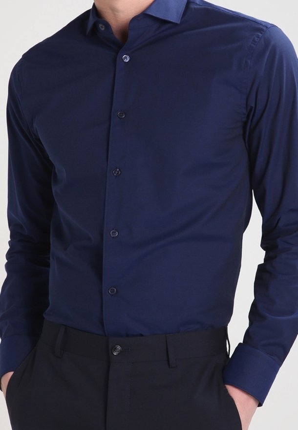 Man in een aansluitend, lang mouw, donkerblauw overhemd met knopen dat in zwarte broek is gestopt, met handen in de zakken.