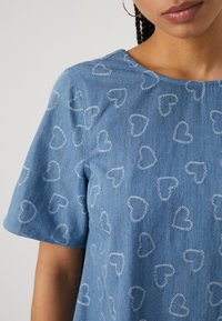 Ανοιχτού μπλέ τζιν πουκάμισο με κοντά μανίκια, διακοσμημένο με λευκές ραμμένες καρδιές σε όλη την επιφάνεια και στρογγυλή λαιμόκοψη.