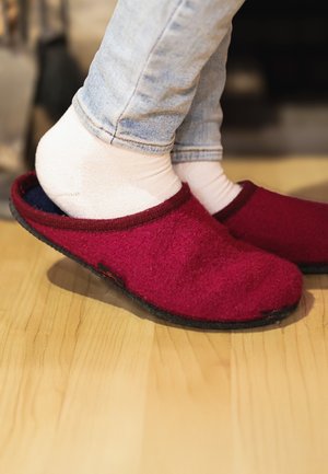 Voeten in witte sokken en lichtblauwe spijkerbroeken schuiven in rode binnenslippers op een houten vloer.