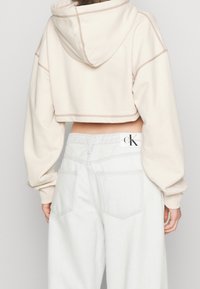Felpa corta beige con cappuccio e polsini a coste, abbinata a jeans azzurri a vita alta con logo Calvin Klein sul retro.
