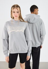 Felpa oversize grigia chiara con grafica frontale, polsini e orlo a coste. Modello che indossa pantaloni corti da biker neri; dettagli di cucitura sottili visibili.