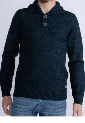Mann trägt einen dunkelblauen Strickpullover mit Zweiknopfkragen und hellblaue Jeans, steht vor einem einfarbigen hellen Hintergrund.