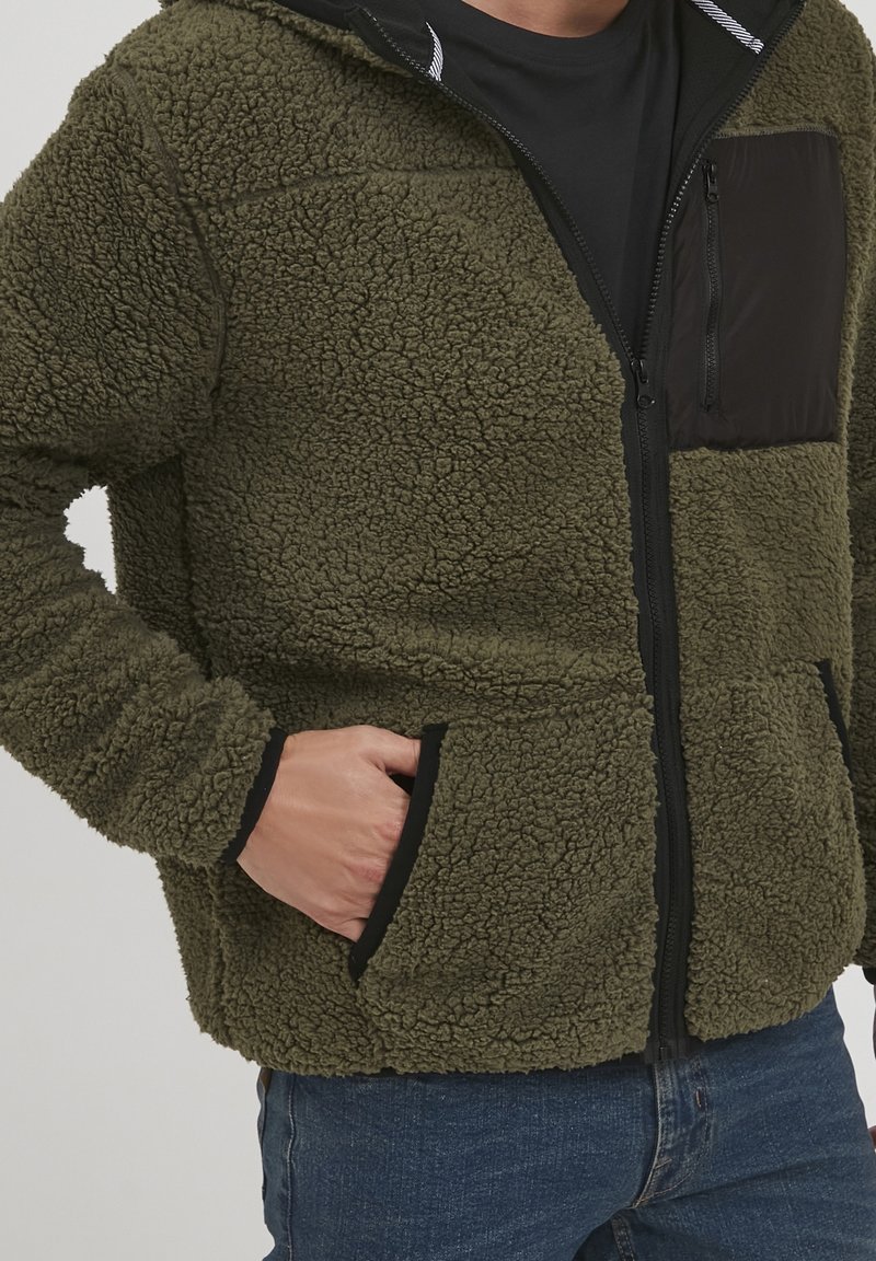 Olijfgroen fleece jack met een gestructureerde stof, rits aan de voorkant en een zwarte borstzak. Voorzien van zijzakken en zwarte accenten.
