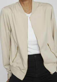 Personne portant une veste aviateur beige zippée sur une chemise blanche et un pantalon noir, assise devant un fond uni.