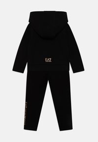 Μαυρό σετ φόρμας που περιλαμβάνει ένα φούτερ με καγκουρό τσέπη και αντίστοιχο παντελόνι. Η χρυσή επωνυμία EA7 προσθέτει αντίθεση και στα δύο κομμάτια.