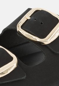 Sandalo in suede nero con due grandi fibbie dorate lucide, design aperto sulla punta e soletta in morbido tessuto.