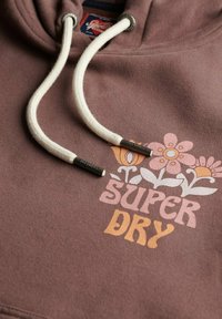 Hoodie marron avec cordons blancs, arborant un design floral et le texte "SUPER DRY" en rose et orange. Tissu doux et texturé.