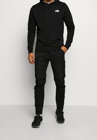 Mężczyzna ubrany w czarną bluzę z kapturem The North Face, czarne spodnie cargo i czarne sneakersy, stojący na jasnym, jednolitym tle.