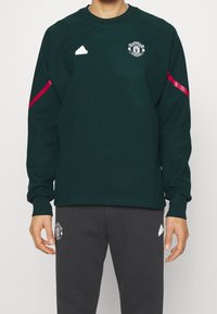 Grön sweatshirt med långa ärmar, Manchester United-logotyp, rosa randdetalj på ärmarna, ihop med mörkgrå byxor, slät textur.