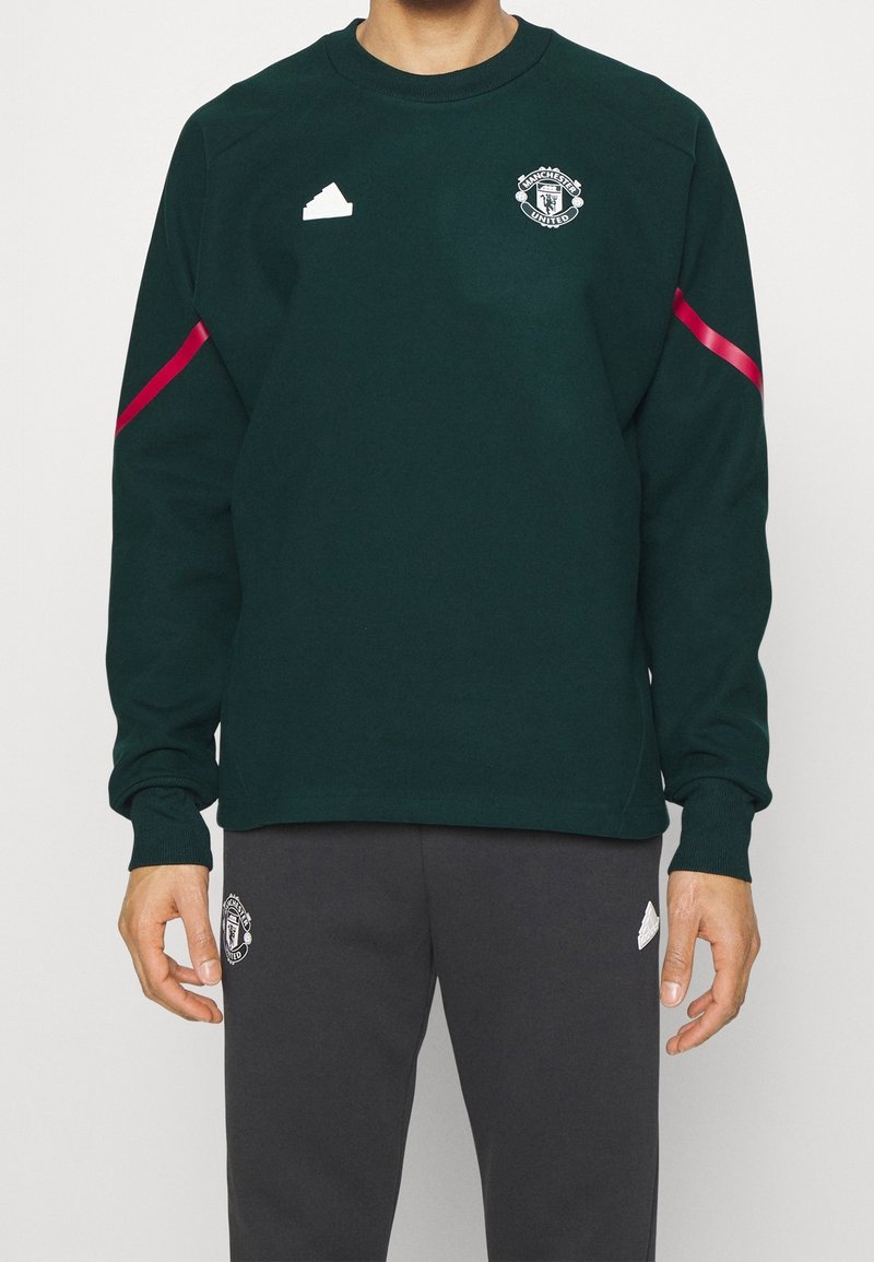 Grön sweatshirt med långa ärmar, Manchester United-logotyp, rosa randdetalj på ärmarna, ihop med mörkgrå byxor, slät textur.