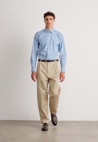 Camisa azul clara com botões, calças beje sob medida, cinto preto e sapatos castanhos. A camisa apresenta um colarinho clássico e um design ajustado.