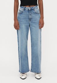 Φαρδιά μπλε τζιν παντελόνια με ψηλή μέση, διαθέτουν μπροστινές τσέπες και ξεθωριασμένο ύφασμα. Συνοδεύονται από ένα ριμπ φόρεμα μαύρου χρώματος και λευκά παπούτσια.