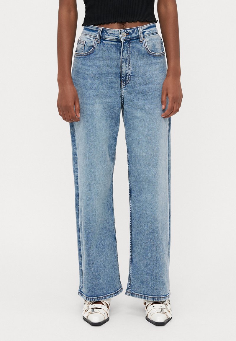 Φαρδιά μπλε τζιν παντελόνια με ψηλή μέση, διαθέτουν μπροστινές τσέπες και ξεθωριασμένο ύφασμα. Συνοδεύονται από ένα ριμπ φόρεμα μαύρου χρώματος και λευκά παπούτσια.