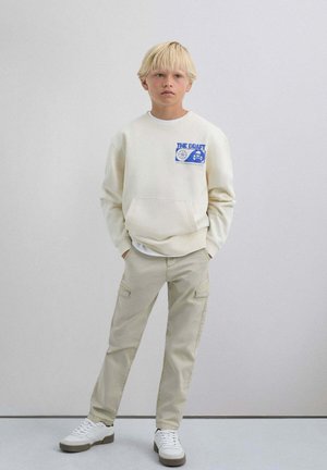 Sudadera crema con diseño impreso en azul, bolsillo frontal; pantalones cargo beige con bolsillos laterales; zapatillas blancas con suelas gris claro.