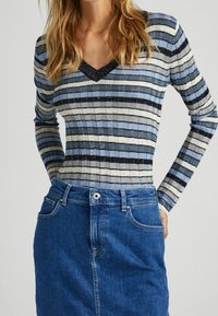 Sweter w pasiaki w odcieniach niebieskiego i szarości, z dekoltem w kształcie litery V i prążkowaną teksturą, zestawiony z niebieską spódnicą dżinsową z kieszeniami.