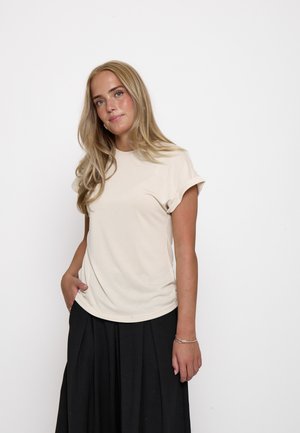 Ung kvinde med langt blondt hår iført en beige kortærmet top og sort plisseret nederdel, står med en hånd i lommen mod hvid baggrund.