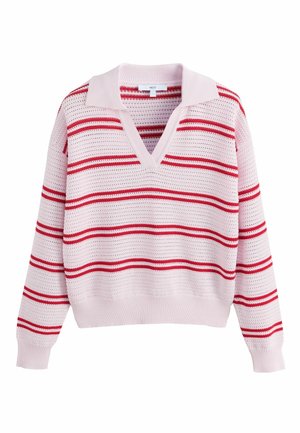 Blond kvinde står med armene over kors iført en hvid og rødstribet sweater, olivengrønne cargobukser og beige kile-sandaler.