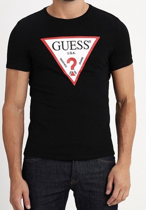 T-shirt noir à manches courtes avec un logo triangulaire rouge et blanc présentant le texte "GUESS U.S.A." et un point d'interrogation, porté avec un jean foncé.
