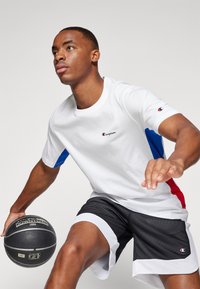 Camiseta blanca con mangas azules y rojas, con un pequeño logo. Pantalones cortos de malla negros con acentos blancos. Sosteniendo un balón de baloncesto.
