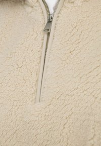 Veste en polaire couleur crème avec une surface texturée, dotée d'une fermeture éclair en métal et d'un bord beige contrastant le long de la fermeture.