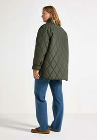 Una persona con capelli lunghi indossa una giacca imbottita verde scuro, jeans blu e scarpe marroni, in piedi davanti a uno sfondo bianco semplice.