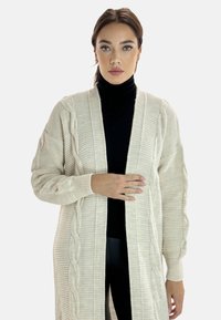 Beige stickad cardigan med kabelmönster, öppen fram, långa ärmar och ribbade ärmslut. Bärs över en svart polo. Slät textur.