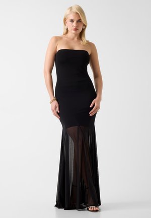 Blonde vrouw in een strapless zwarte jurk met doorzichtige rok, staand tegen een witte achtergrond, gecombineerd met een gouden armband en oorbellen.