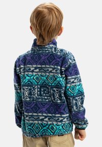 Pull en polaire avec des motifs géométriques violets, bleus et turquoises. Col montant et tissu texturé avec une coupe décontractée.