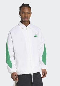 Giacca bianca con dettagli verdi, chiusura con zip, maniche raglan e polsini elasticizzati; presenta un piccolo logo Adidas verde sul petto.