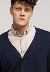 Granatowy sweter z dzianiny o głębokim dekolcie w kształcie litery V, z zapięciem na guziki i teksturowanym materiałem. Nałożony na jasnobeżową koszulę z guzikiem.