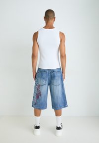 Denim-Shorts mit einem verblichenen blauen Waschung, mit roter Stickerei auf der linken Seite und zwei Gesäßtaschen. Entsprechendes weißes Tanktop sichtbar.