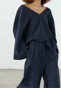 Femme portant un haut ample à fines rayures sombres en V et un pantalon taille haute assorti avec des poches sur un fond uni.