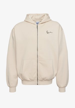 Krém színű cipzáros kapucnis pulóver elülső zsebből, bordázott mandzsettából és diszkrét hímzett logóból áll. Puha, texturált anyag és laza fit.