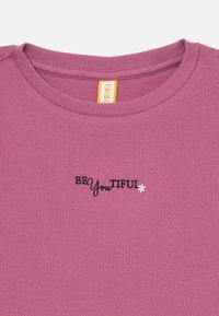 Lilla ribbet t-skjorte med svart brodert tekst "BE YouTIFUL" med en liten hvit blomst som akcent. Rund halsdesign med synlig søm.