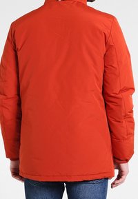 Červená izolační bunda s vysokým límcem, prošívanou texturou a bočními kapsami. Má žebrované manžety a hladký povrch látky.