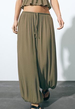 Pantaloni largi, verzi măsliniu, cu talie cu șnur și manșete elastice, purtați cu sandale negre pe un podea gri.