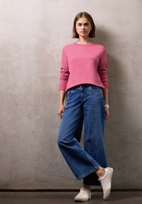 Donna in piedi contro un muro di cemento, che indossa un maglione rosa, jeans a gamba larga blu, sneakers bianche e braccialetti d'argento al polso.