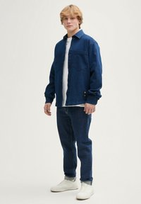 Jeansowa kurtka w ciemnoniebieskim kolorze, z długimi rękawami i zapięciem na guziki, w zestawie z dopasowanymi dżinsami i białymi trampkami. Prosty design i luźny krój.