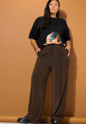 Una modelo posa sobre un fondo tostado, con una camiseta negra con estampado gráfico, unos pantalones marrones brillantes de pernera ancha y unos zapatos negros robustos.